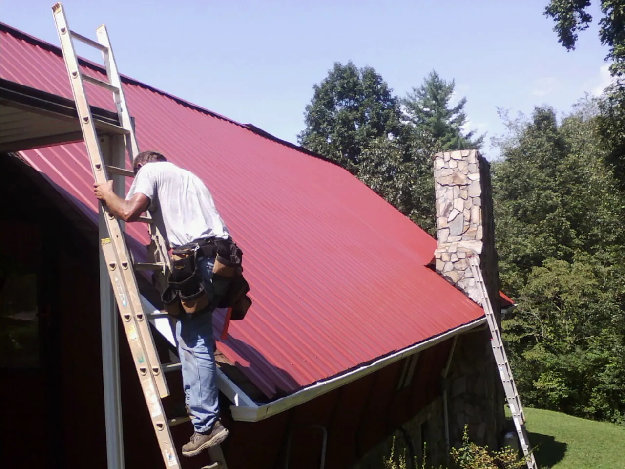 Roofer on a ladder beside a red metal roof and stone chimney — Soffit & Fascia Repair by Roofing Brothers in Ladoga
