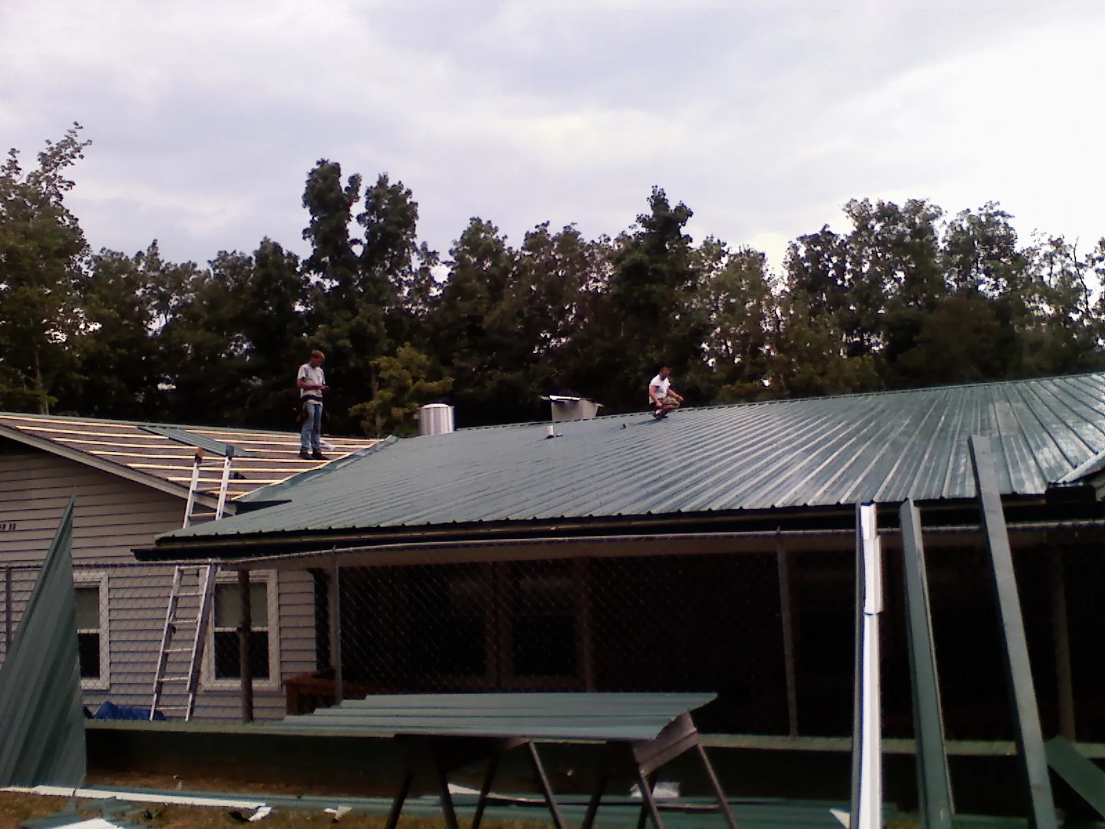 Two roofers installing green metal panels above a covered porch in Ladoga